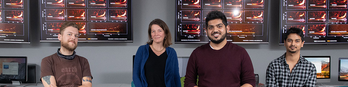 Center for Relativistic Astrophysics Director and School of Physics professor Laura Cadonati (second from left) with members and graduates of the Georgia Tech LIGO research team (from left) James Alexander Clark, Karan Jani, and Sudarshan Ghonge. Behind them, a chart shows gravitational wave signals from cosmic events measured by LIGO-Virgo. (Credit: Allison Carter)